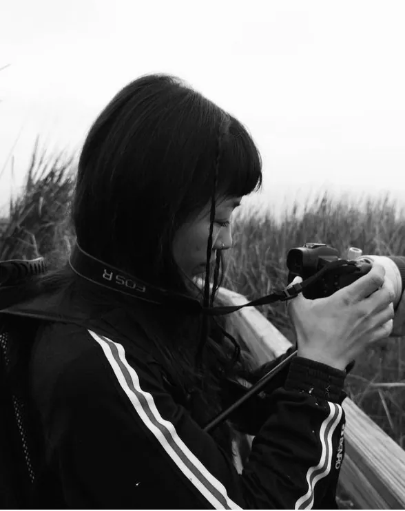 Retrato en blanco y negro de Alexandra Pinto mirando el visor de su cámara, de perfil, en un paisaje de pastizales.