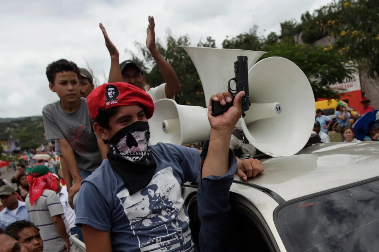 Foto de Edgard Garrido: un joven con boina roja del Che y pañuelo de calavera levanta una pistola junto a parlantes sobre un auto, en medio de una manifestación.