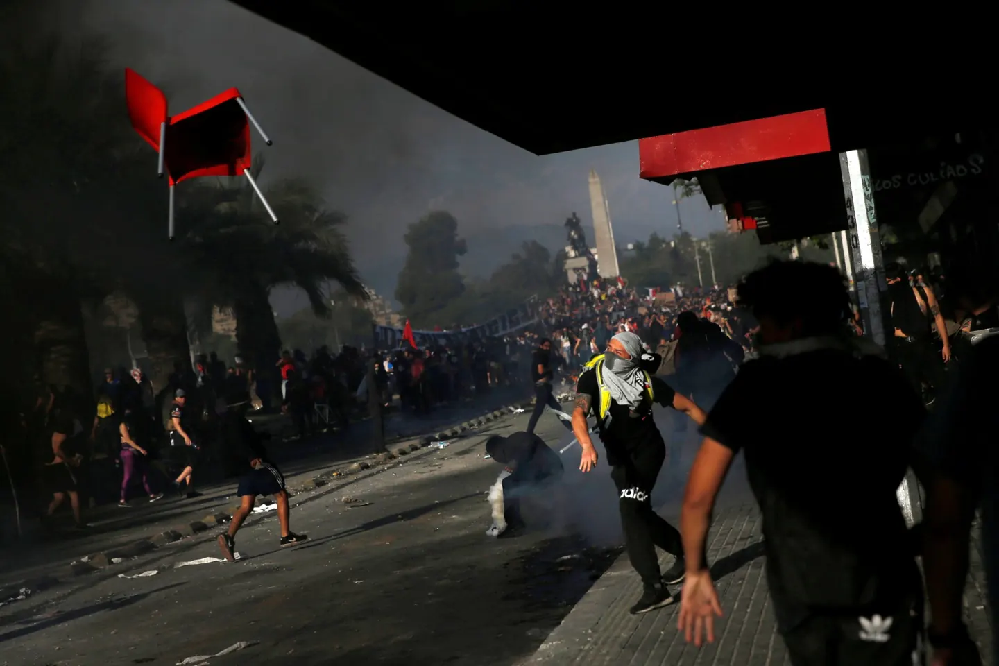 Foto de Edgard Garrido: manifestantes entre el humo bajo un alero, una silla roja vuela por el aire y al fondo la multitud rodea el monumento de Plaza Baquedano.