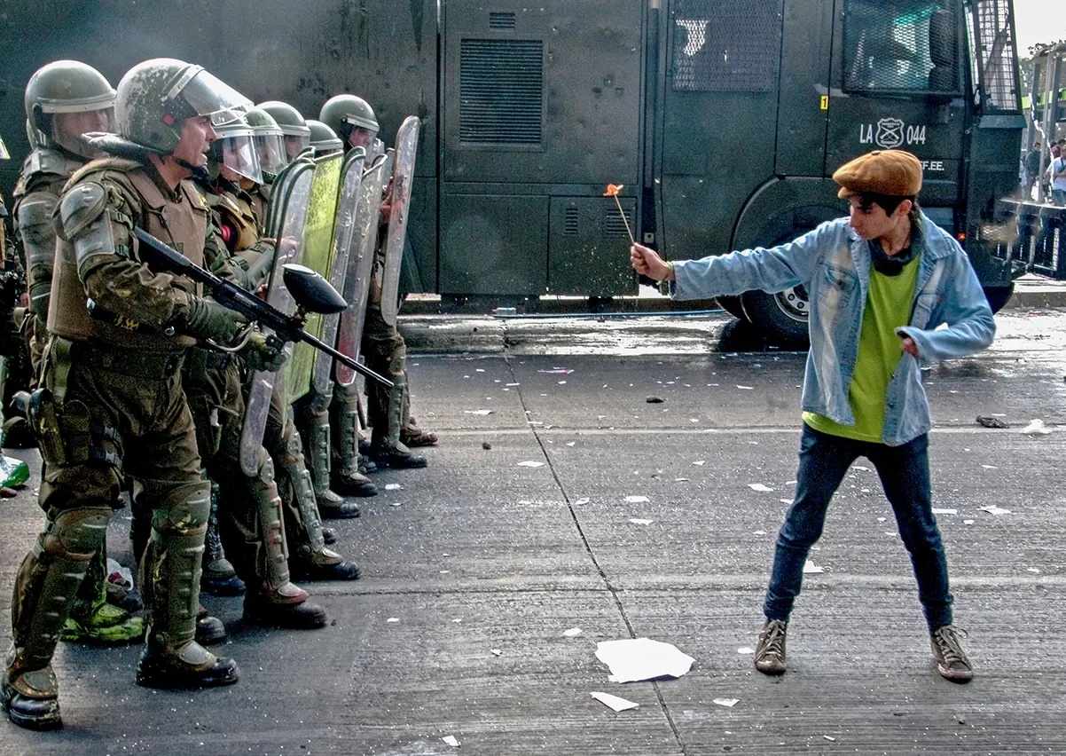 Foto de Héctor González de Cunco: un joven con boina y chaqueta de mezclilla ofrece una flor a una fila de carabineros de fuerzas especiales con escudos, frente a un carro policial.