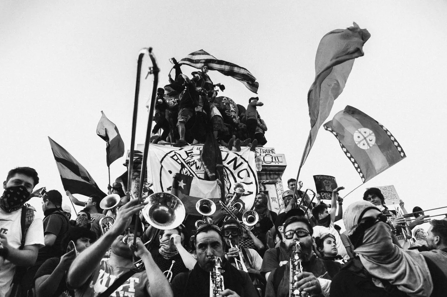 Foto de Alexandra Pinto: una banda de bronces toca entre la multitud al pie de un monumento cubierto de manifestantes con banderas, entre ellas la wenufoye.