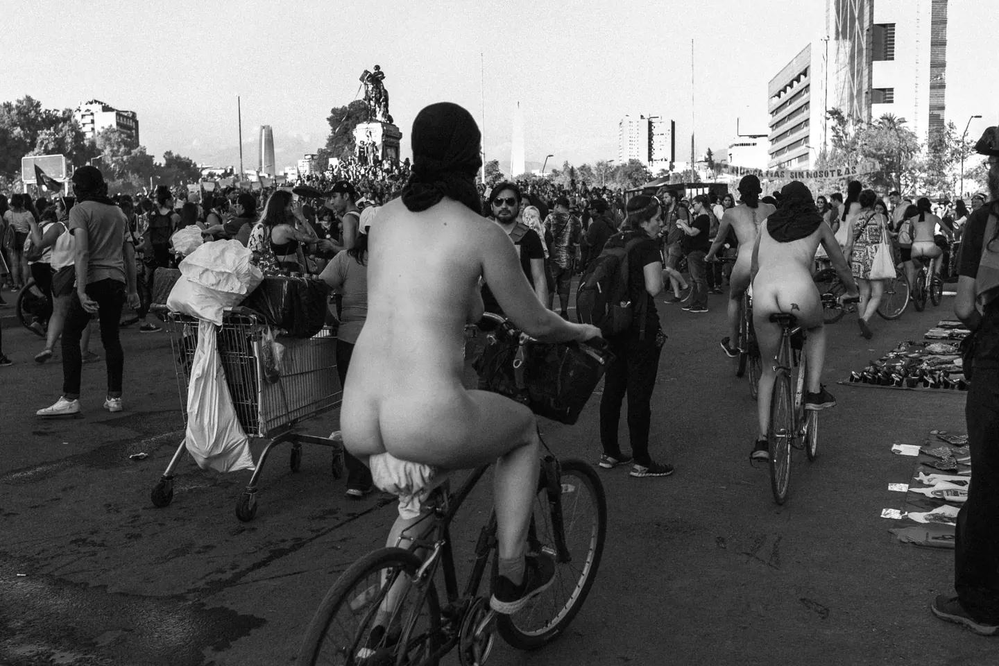 Foto de Alexandra Pinto: ciclistas desnudas con el rostro cubierto avanzan en bicicleta entre la multitud de una marcha, con el monumento de Plaza Baquedano al fondo.
