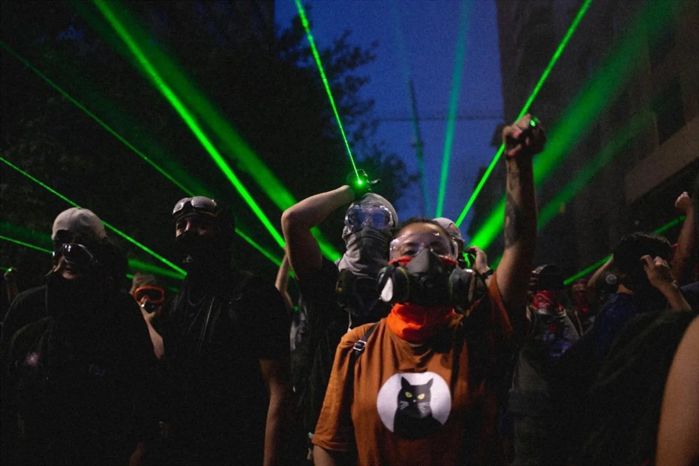 Foto de Alexandra Pinto: manifestantes con antiparras y máscaras antigás, de noche, bajo rayos de láser verde; una mujer levanta el puño.